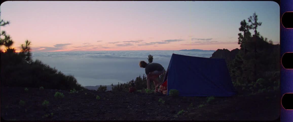 Wide shot of a man climbing out of his tent and stretching in front of mountain sunrise scenery above the clouds