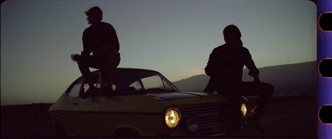 Two silhouettes sitting on a yellow car in front of a dusty mountain desert scenery after sunset