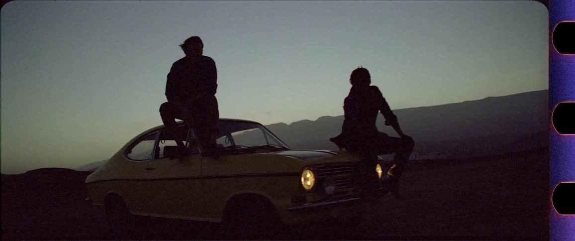 Two silhouettes sitting on a yellow car in front of a mountain desert region after sunset