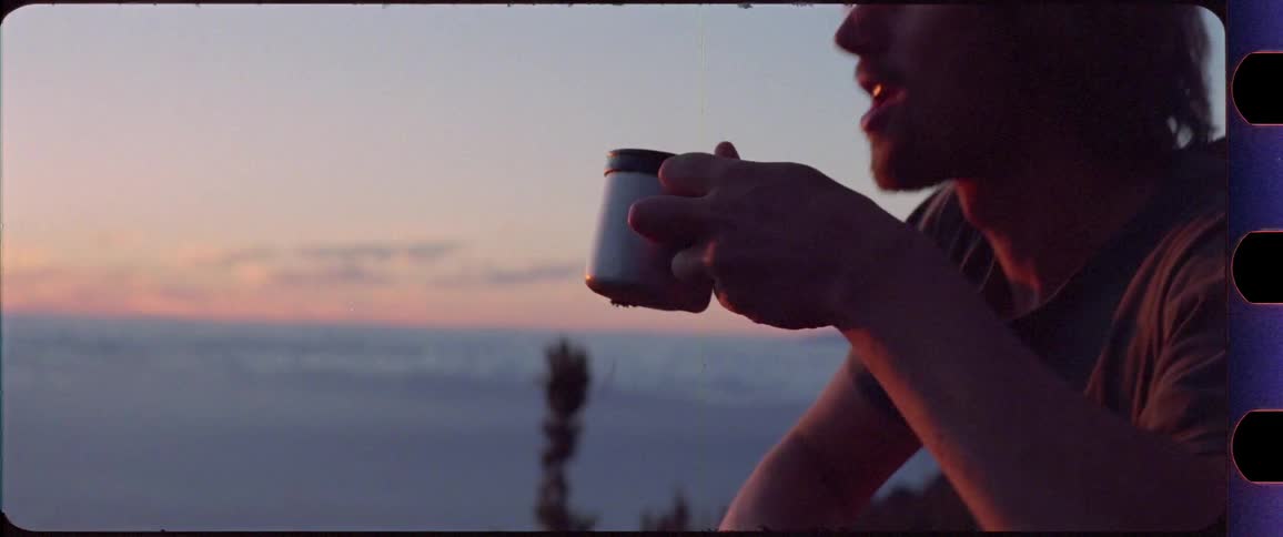 Man pouring coffee from a pot to his cup and then drinking it while overlooking sunrise on top of a mountain