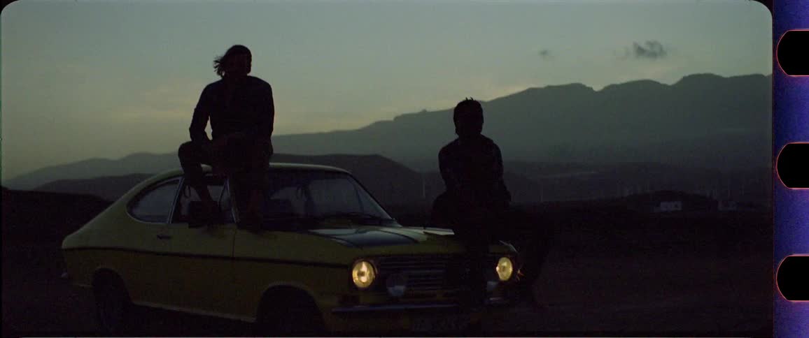 Two silhouettes sitting on a yellow car in front of a mountain desert region after sunset