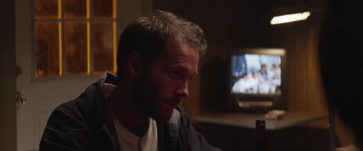 A man is sitting down with a bottle in front of him and a television on in the background