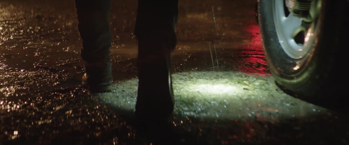 A person in boots carrying a flashlight walks through the mud in the rain