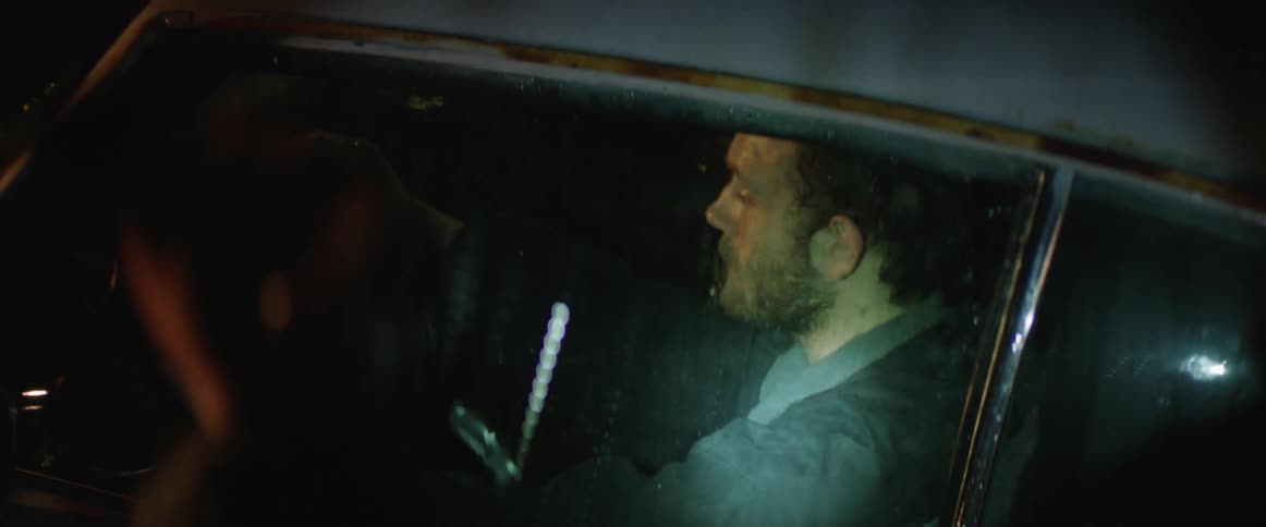 On a rainy night, a man with a flashlight approaches a car where a man is seated