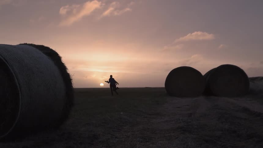 Two people run through a field with hay bales at sunset