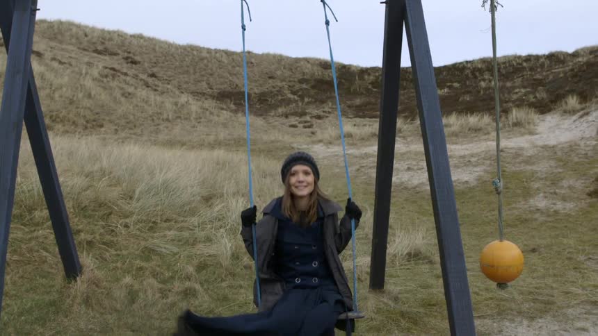 A woman wearing a hat and scarf swings on a swing set on a windy day