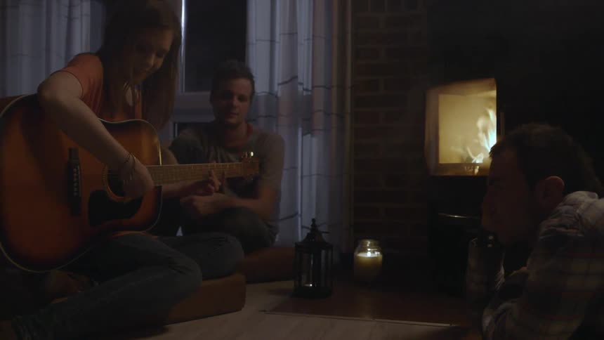A woman plays guitar by the fireplace as people sing along