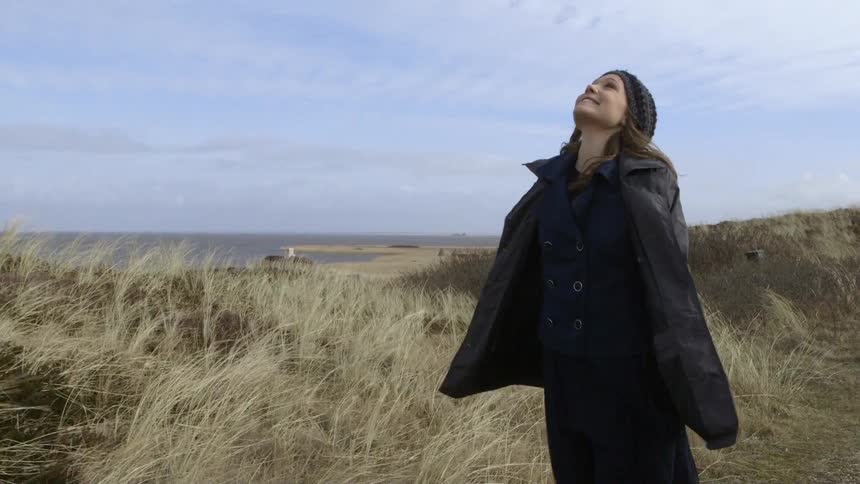 A woman in a coat walks along a path above a beach on a windy day