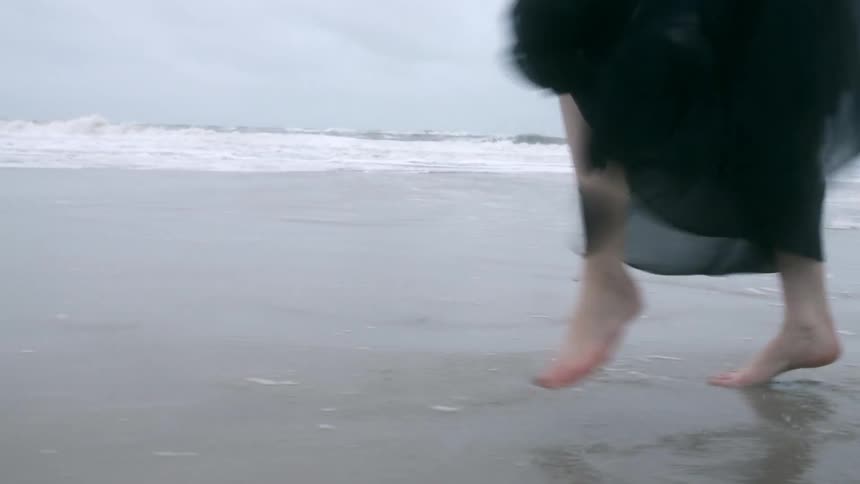 A woman in a black flowy dress skips and dances along the beach