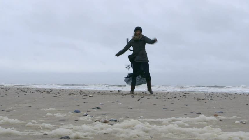 A woman in a winter hat and scarf twirls and dances on the beach
