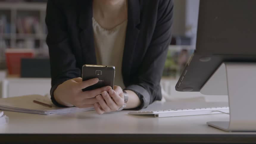 A woman is sitting at a desk typing and using a cell phone