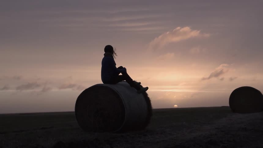 A woman sits on a hay pile as she watches the sun set