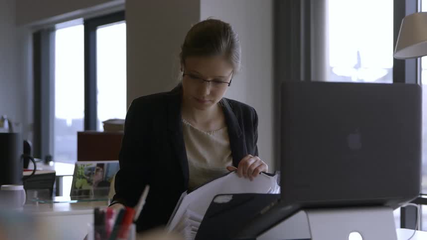 A woman wearing glasses is putting papers away by a computer