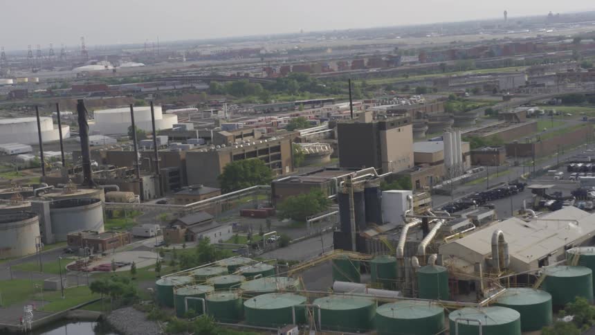 Cars are parked in lots around factory buildings with smoke stacks in an industrial district