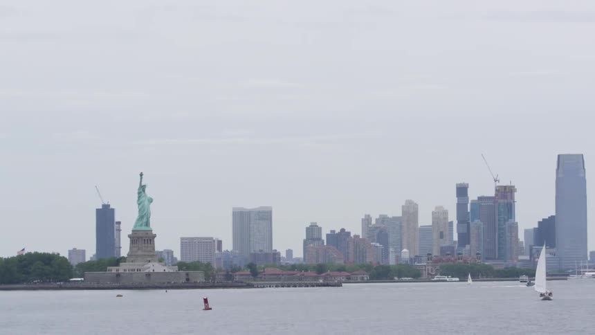 The statue of liberty stands on its island across from towering metropolitan area as a few small boats sail by