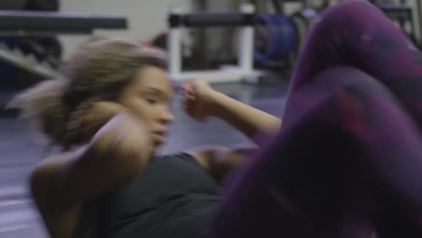 A young woman is on her back on a padded floor mat in a gymnasium where she is doing a very vigorous abdominal exercise