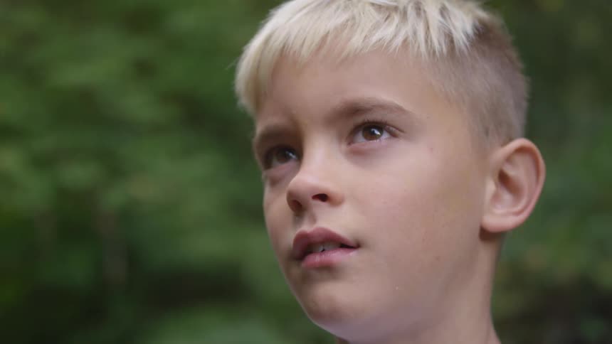A blond boy looks up at the sky with trees behind him