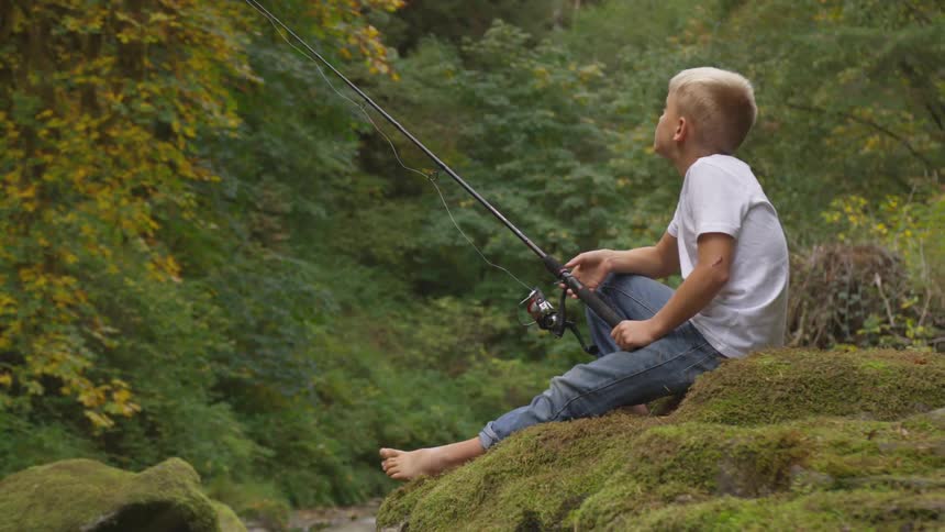 A young boy is holding a fishing rod on a mossy ledge while looking up into the trees