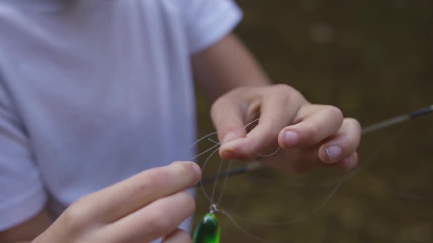A youngster in a t shirt is carefully tying a lure to the fishing line on their fishing rod