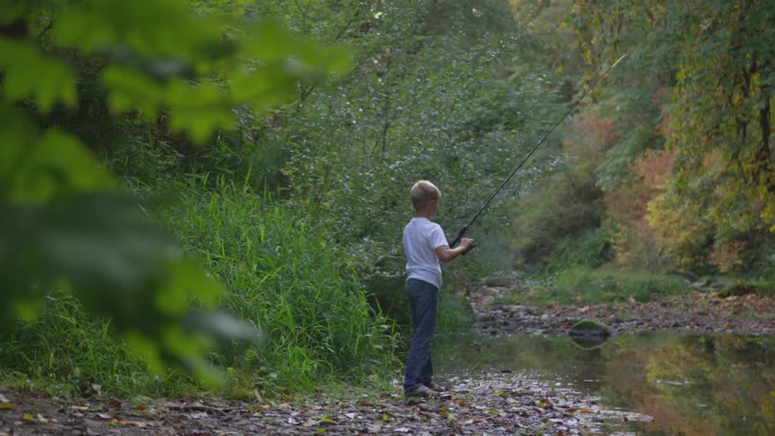 Boy reels in fishing line on shore of stream during the day