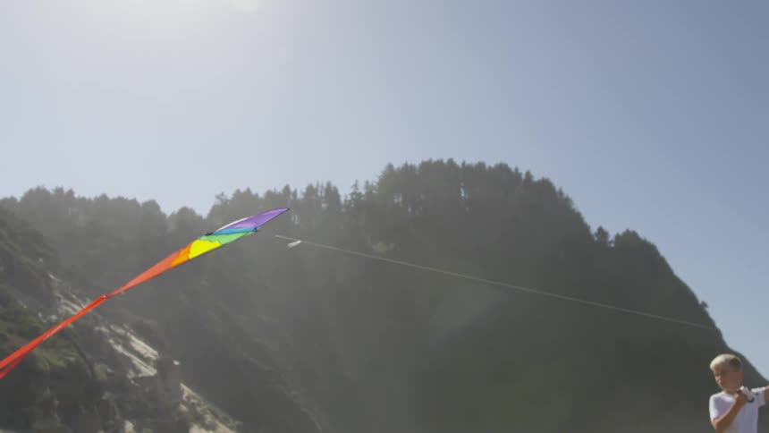 A boy holds a kite string up high as he flies his colorful kite on the beach