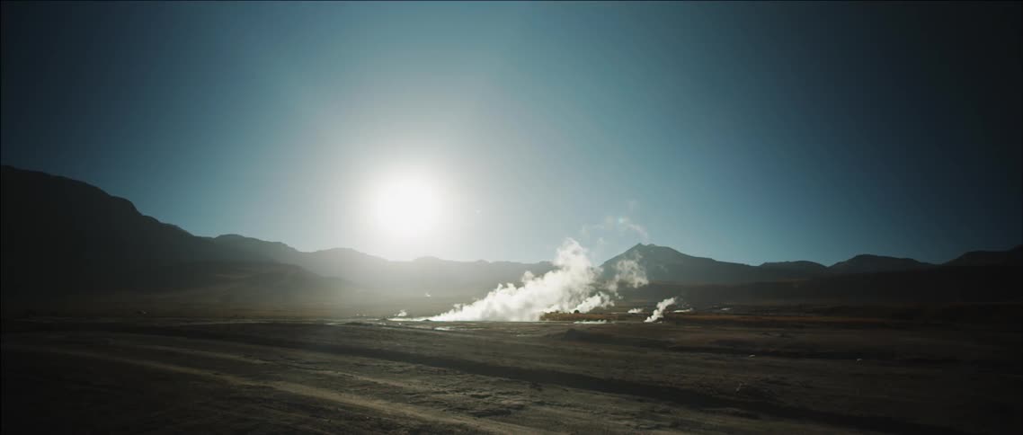 Steam rising from a geyser in a flat valley with a rising sun in the background