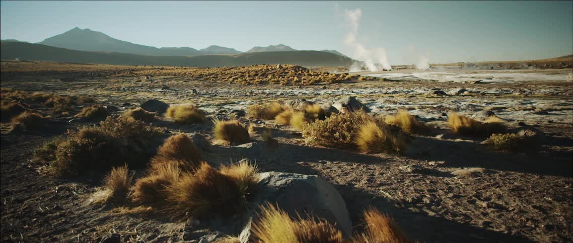 A large field filled with rocks and sand has smoke rising in the distance