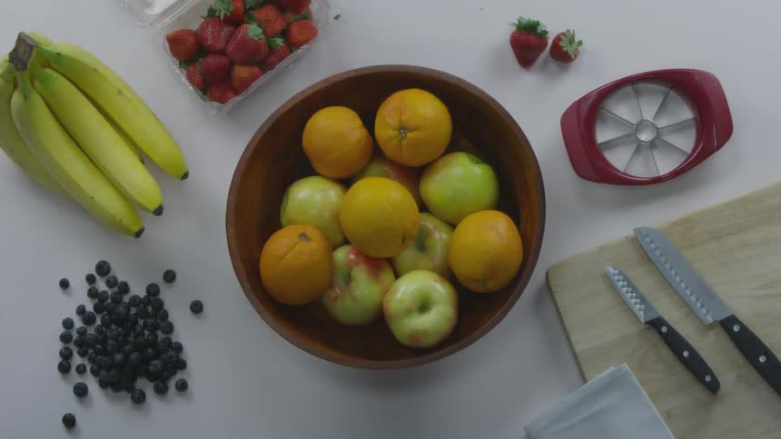 Someone puts an orange into a bowl of fruit on a prep table filled with fruit