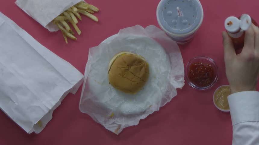 A white person sitting at a table with food on it and pouring ketchup in a bowl before dipping a fry in the ketchup