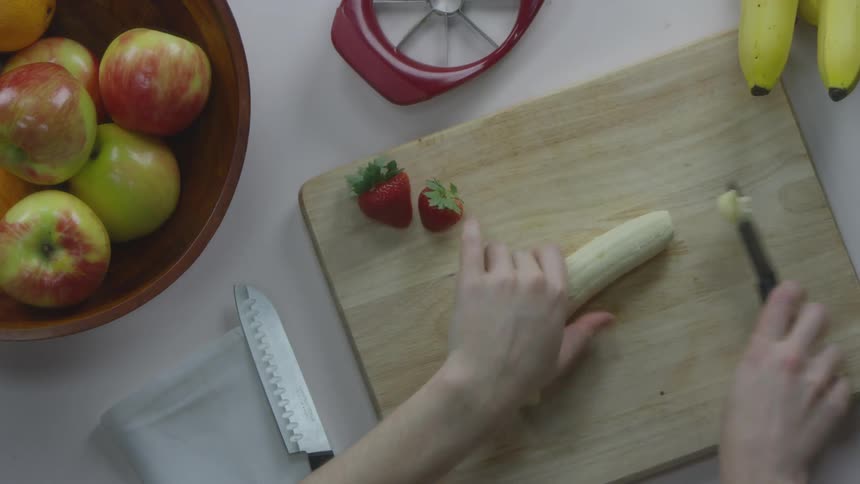 A woman's slender hands pick up a small knife and then begins to carefully chop a peeled banana into thin slices on a wooden cutting board
