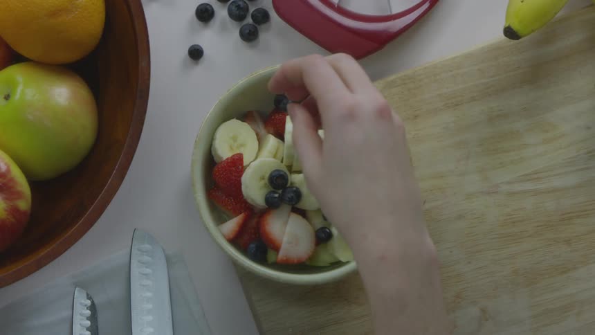 A young man picks up blueberries and adds them to a bowl of sliced bananas and strawberries