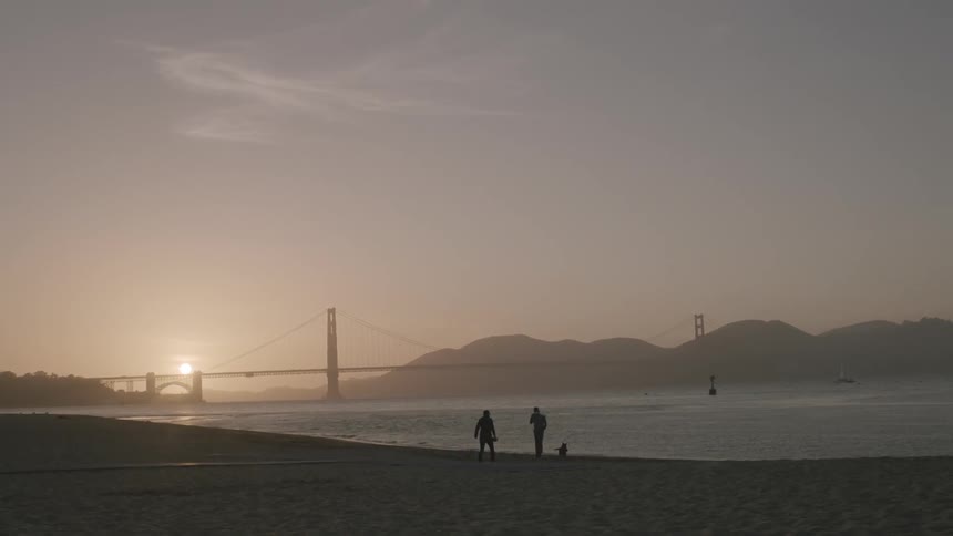 Two people and a dog walk on a beach at sunset with the golden gate bridge in the background