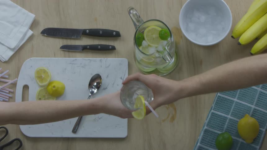 Above a kitchen work space with a cutting board one persons hand is seen handing another person a glass full of a clear liquid with ice and a lemon slice on the side