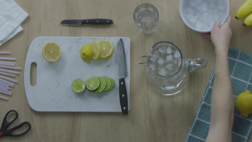 A person puts ice into a pitcher of water then adds cut lemons and limes