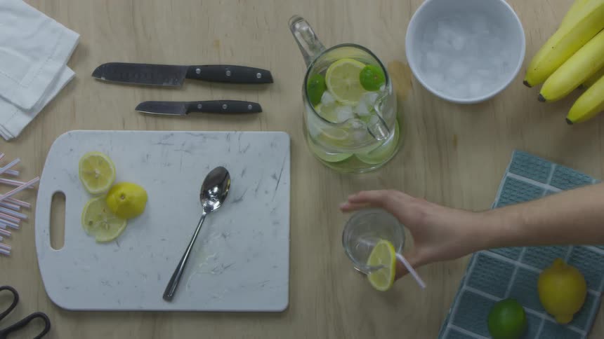 A hand pits a glass on the table where there is a cutting board with cut lemons knives and a pitcher of ice water with lemons already added