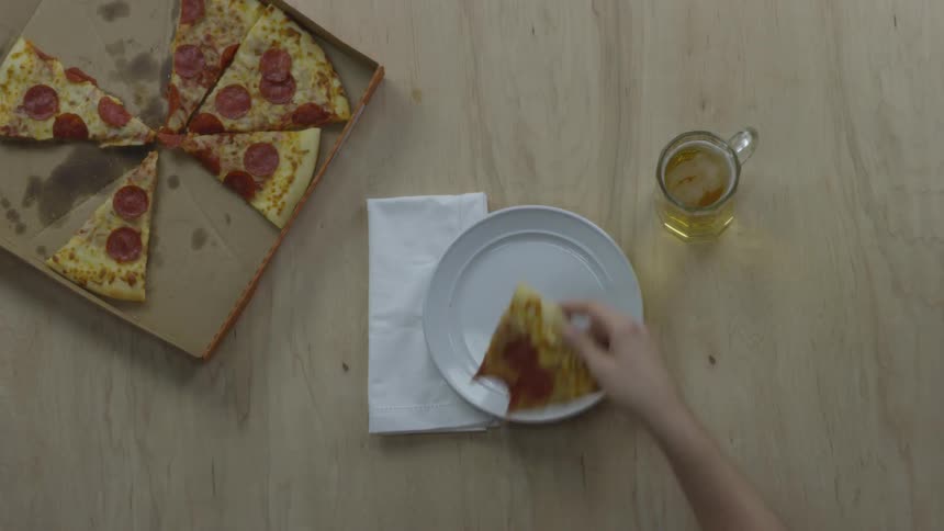 A partly eaten pizza is on the table with a white plate and a hand is placing a piece of pizza on the plate with a missing bite