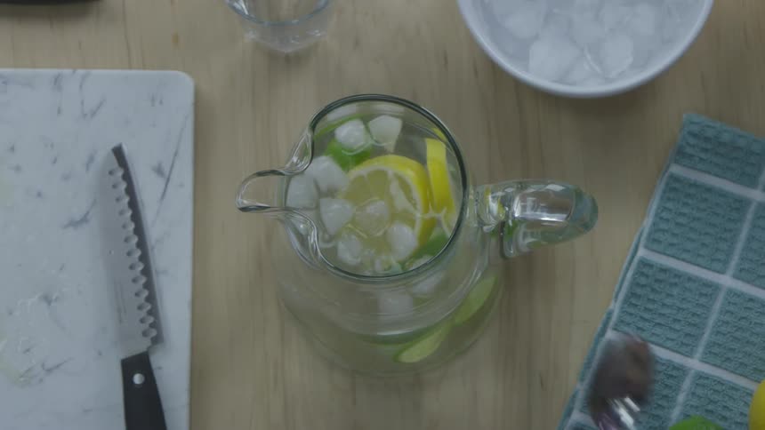 A person stirs a water pitcher with ice cubes and lemon and lime slices in it