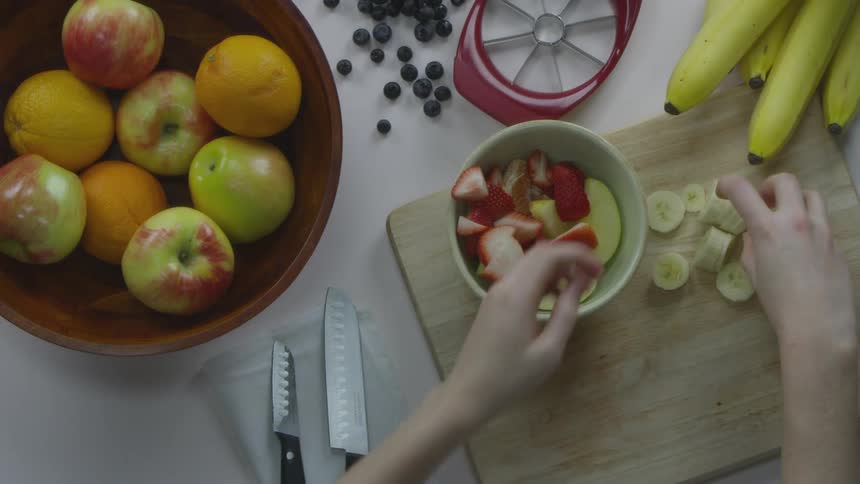 A white person placing a ceramic bowl on a wooden cutting board and filling the bowl with sliced fruits