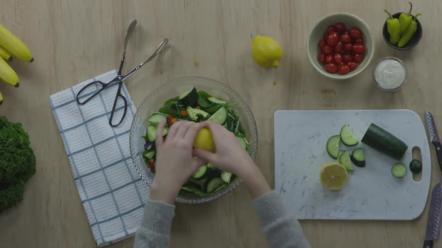 A person cuts a lemon in half and squeezes it over a salad in a glass bowl