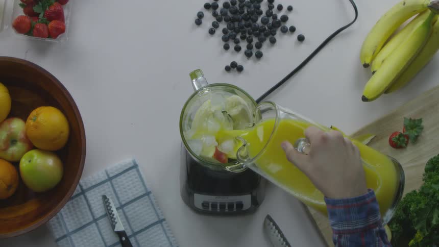 A person lifting a pitcher of juice from a table covered in fruits and pouring it into a blender containing sliced fruit