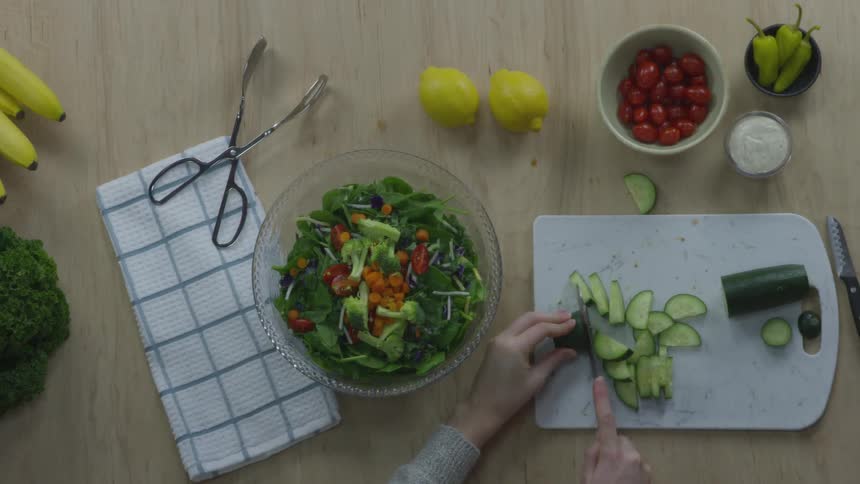 Next to a colorful bowl of salad and woman carefully slices a cucumber on a cutting board and then scoops the pieces and places them in the bowl with the salad