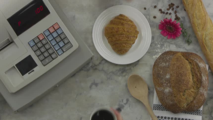 A person presses a few keys on a cash register and then hands a cup of coffee to another person over a countertop that has some bread and pastries sitting on it as well