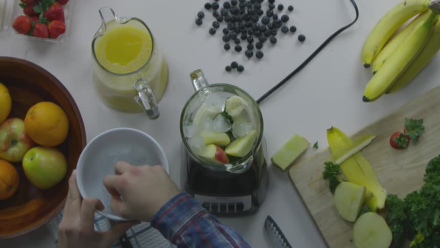 Person takes fruit from bowl and places it in blender which sits on a prep table full of fruit and vegetables