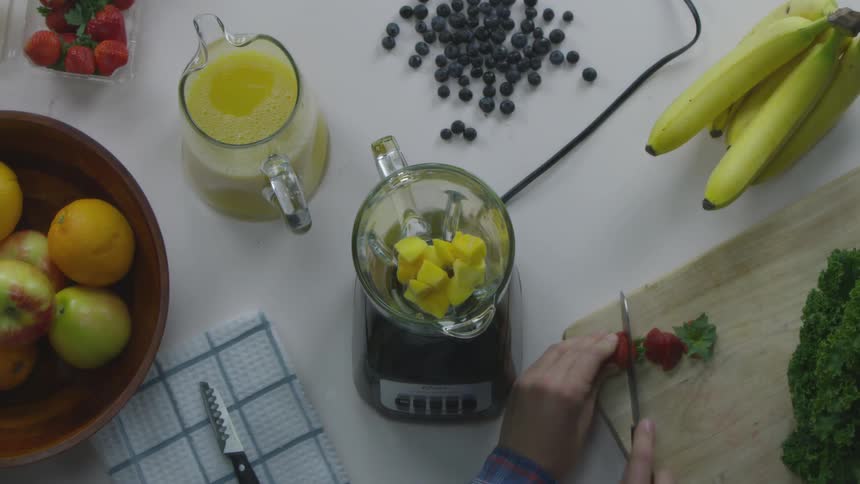 A strawberry is sliced on a cutting board and added to a blender with other fruit