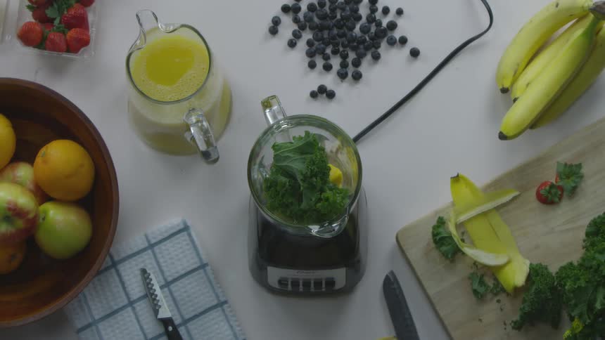 A man works on a kitchen counter surrounded by fruit and juice and peels a banana and adds it to a blender with kale