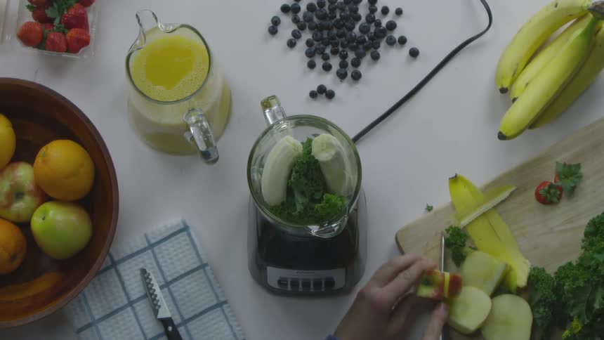 A man picks an apple out of a bowl and slices it on a cutting board before placing it into the blender