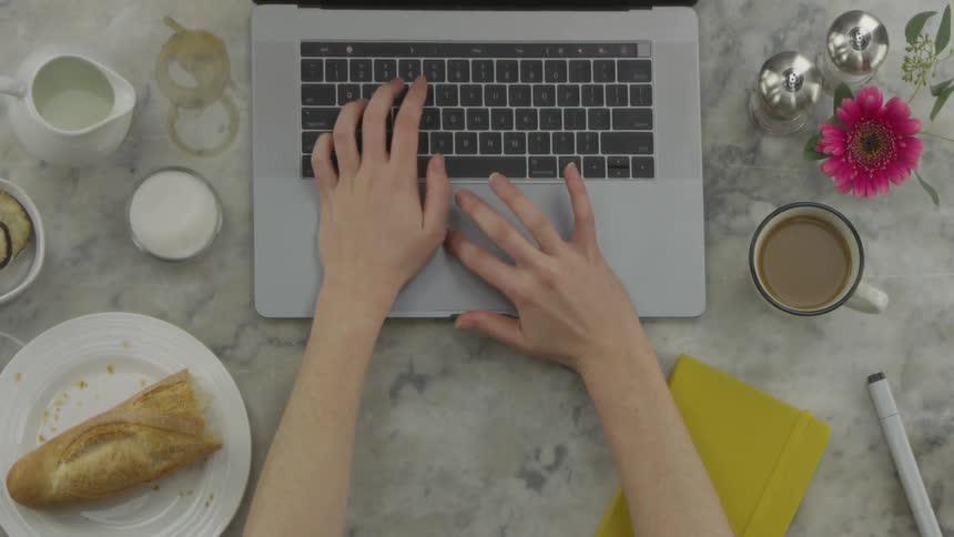 A woman works on her laptop on a coffee stained restaurant counter with food and coffee around her