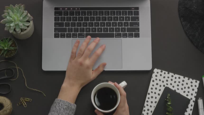 A person holding a cup of coffee taps on the touchpad of a laptop on their desk