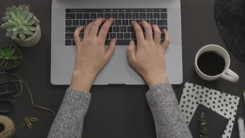 While sitting at a desk with small potted plants and a cup of coffee the slender hands of a young woman are seen typing and then closing the laptop