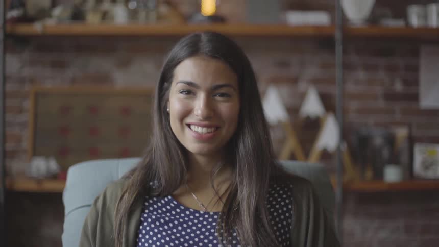 A dark haired female in a polka dot top sites in a chair smiling in front of a brick wall with shelves
